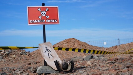 Red danger sign with skull and crossbones and caution tape cordoning off barren land, symbolizing minefield hazard, unexploded ordnance risk and urgent need for demining efforts © Canary4stock