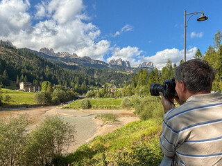 A photographer stands on a ridge capturing a stunning Trentino Alpine valley, with a winding river and a serene lake glowing under crisp mountain light.