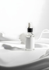 A clear glass dropper bottle with a white label sits on a white silk fabric.