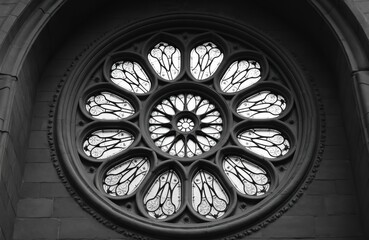 Monochrome gothic rose window of church. Geometric patterns form ornate circular design. Stone architecture frames the stained glass detail. Light shines through ornate glass panes.