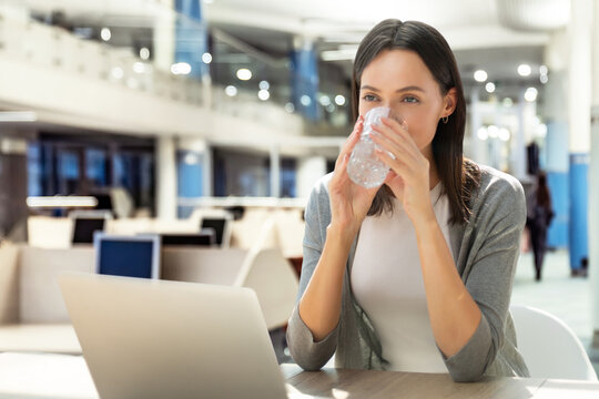 Positive caucasian businesswoman enjoying a refreshing break while drinking a glass of water, sitting at her desk in a modern office with a laptop