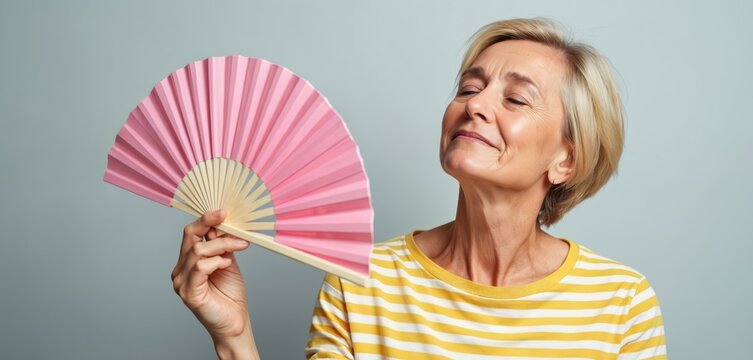 Blonde middle-aged woman fans with pink fan. Closes eyes, finds comfort from hot flash. Older female cools down, feeling relieved. Mature lady experiences menopause symptoms, seeking ease. Face shows