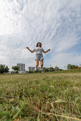 Young woman with dark hair wearing casual clothing jumps joyfully in a grassy field under a bright blue sky, capturing a moment of happiness and freedom in nature