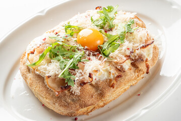 A close-up, high-angle studio shot of an oval flatbread (pinsa or breakfast pizza) topped with arugula, cheese, possibly meat, and a runny egg yolk on a white plate
