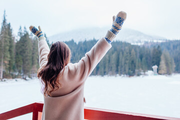 Back view of woman tourist admiring snowy mountain Synevyr lake view wearing white warm fur coat. Winter landscape