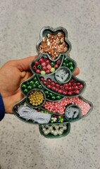 Handheld Christmas tree-shaped container organizes various colorful crafting beads. Compartments are filled with metallic copper stars, red and green spherical beads, gold and silver beads, decor