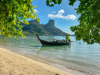 A solitary traditional long-tail boat is anchored in serene, sandy bay, form perfectly framed by vibrant green leaves of overhanging trees, with limestone mountains creating a picturesque backdrop.