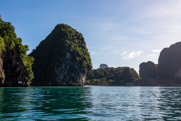 Naklejka premium Majestic limestone karst formations covered in lush green jungle rise from tranquil turquoise Andaman Sea on clear sunny day. Pristine sandy beach is nestled at the base of cliffs in Railay Bay, Krabi