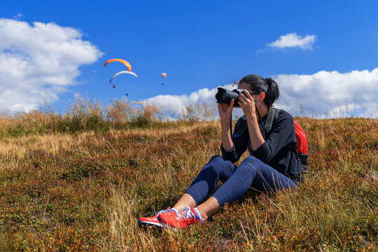A woman wearing a backpack and red shoes sits on a grassy hillside, looking through a professional camera telephoto lens at paragliders floating against a bright blue sky with white clouds.