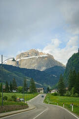 Mountain road view in the Dolomites with a village, green fields and a rocky snow-capped peak in the background.