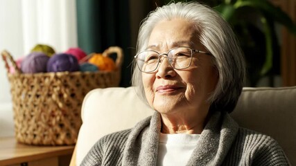 Elderly asian woman with glasses smiling, sitting in a chair with a basket of yarn - Powered by Adobe