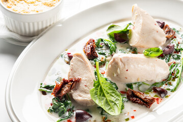 A delicious, creamy Tuscan chicken dish with spinach, sun-dried tomatoes, and olives is served on a white plate with a side dish in a restaurant setting