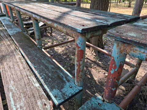 A close-up of a weathered, rustic picnic table and bench with peeling blue and red paint on its rusty metal frame and decaying wooden slats, set outdoors on dirt ground. - Powered by Adobe