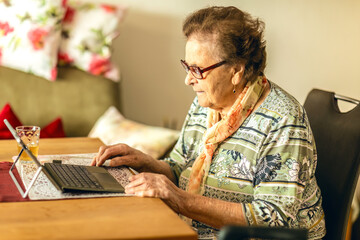 An elderly woman sits indoors and focuses on using a tablet, illuminated by warm natural light.