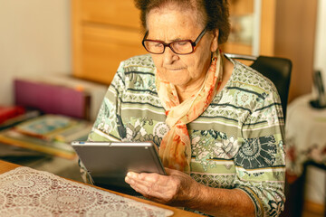 An elderly woman sits indoors and focuses on using a tablet, illuminated by warm natural light.