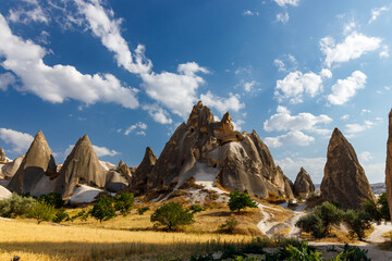 Cappadocia. T&uuml;rkiye. Rock formations under a blue sky with scattered clouds in a dry, grassy landscape. Sparse green trees dot the foreground.