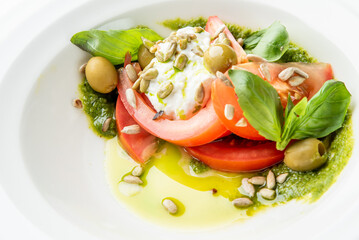 A vibrant close-up of a fresh burrata caprese salad with sliced heirloom tomatoes, fresh basil leaves, green olives, pesto, olive oil, and sunflower seeds in a white bowl.
