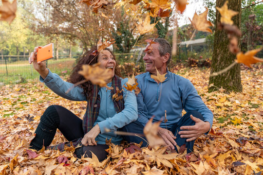 Mature couple sitting on autumn leaves taking a selfie with a smartphone, smiling happily