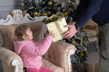 a little girl in a pink sweater is sitting in a chair and being handed a large box wrapped in Christmas paper