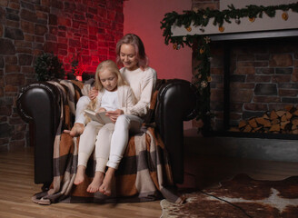 Mom and her daughter are sitting in a chair by the fireplace in the evening,