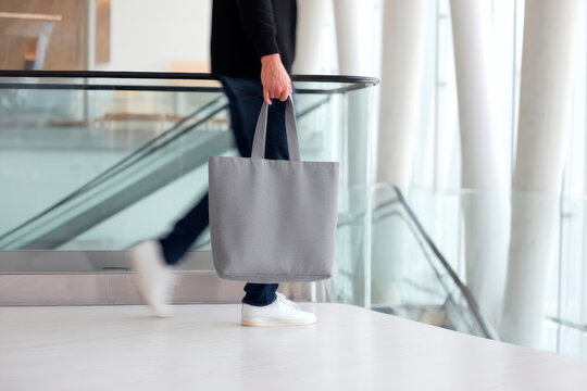 Minimalist grey tote bag mockup shown in a modern indoor space as a man walks forward. Blank fabric surface for branding, logo placement, retail identity, or eco-friendly shopping design. - Powered by Adobe