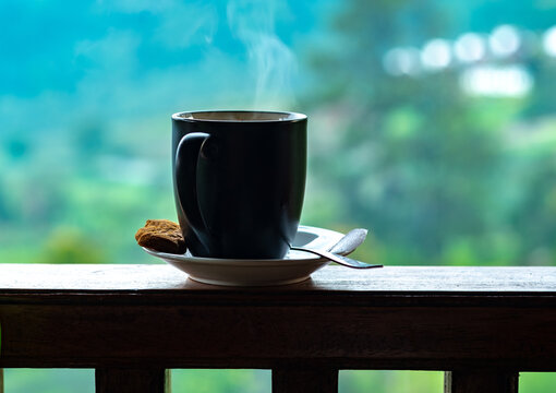 A steaming mug of coffee and a small biscuit resting on a wooden balcony railing, framed by a serene, misty nature backdrop. A perfect, peaceful morning moment.