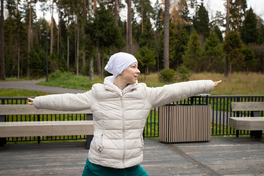 Bald woman after chemotherapy practicing yoga outdoors on a wooden terrace surrounded by forest.