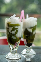 two glasses of refreshing Es Cincau Hijau drinks on a reflective table. Green grass jelly, coconut milk, and shaved ice create a cooling traditional Indonesian treat against a lush blurred background