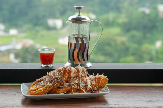 Crispy fried bananas topped with cheese and chocolate, served with French press coffee on a balcony overlooking a lush green valley. A delicious Indonesian snack with a scenic view.