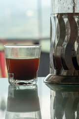 Close-up of black coffee beside a metallic French press on a mirrored table. Sharp reflections and condensation details capture the essence of a fresh, hot brew ready to be enjoyed.