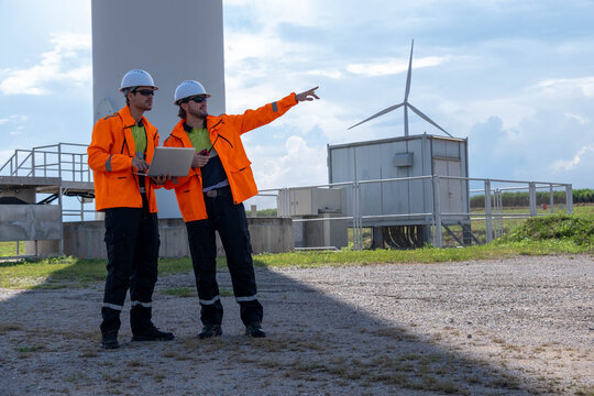 Two workers in bright orange jackets discuss wind turbine operations at a renewable energy site during a sunny day