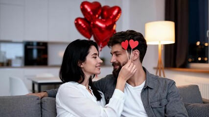 Affectionate couple celebrating a romantic evening at home. A woman playfully holds paper hearts while looking at her partner on the sofa. Valentine's Day and love concept