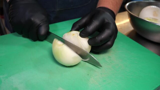 A chef wearing black sanitary gloves using a sharp knife to rapidly dice white onions on a bright green cutting board in a professional kitchen setting. The chopped pieces are then scooped into a meta