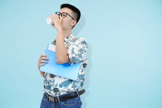 A focused young Asian man wearing glasses and batik shirt, drinking from a disposable coffee cup while reviewing a document held in a blue folder against a light blue studio background