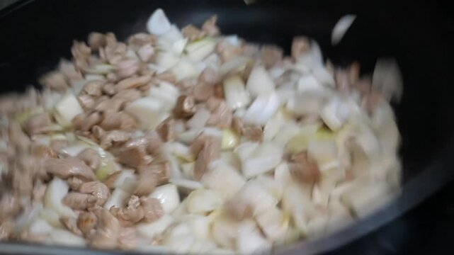 White onions, chopped celery, and a mix of diced carrots and potatoes are consecutively added to and stirred with cooking chicken pieces in a large black pan on the stovetop.