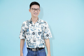 A cheerful young Asian man wearing glasses and batik shirt, standing and smiling confidently while...