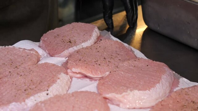 A gloved chef uses a metal shaker to sprinkle ground pepper onto raw, tenderized pork chops arranged on paper towels, then flips and places additional chops onto the board for seasoning in a kitchen.