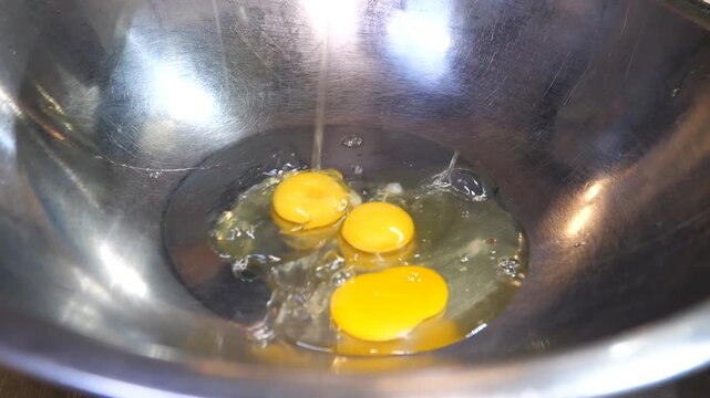 A professional chef wearing black gloves carefully cracks a large number of eggs into a stainless steel bowl, then uses a wire whisk to beat the eggs until they are combined into a smooth