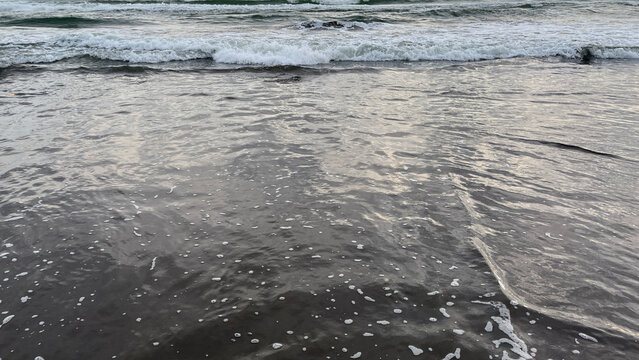 a gentle ocean wave washing over a dark sandy shore, creating foam patterns and textured water. natural beach scene with calm movement and simple composition - Powered by Adobe