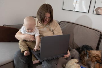 mother holding baby at laptop on sofa, smiling while working on call with dog nearby and cozy livingroom