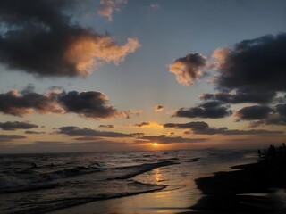 A peaceful sunset over the sea with warm golden light reflecting on the waves and shoreline, surrounded by dramatic clouds that create a calm and atmospheric coastal scene.