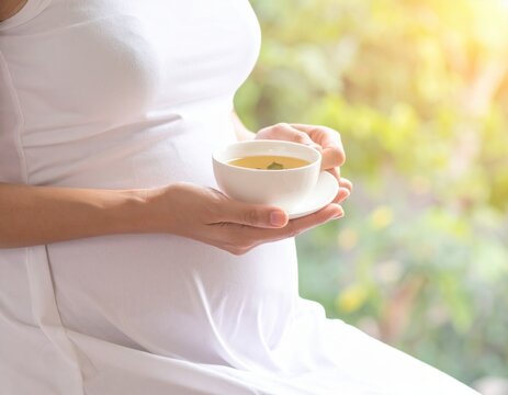 Pregnant woman in white clothing holding cup of herbal tea with natural outdoor bokeh background depicting prenatal wellness - Powered by Adobe