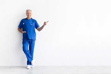 Senior male doctor in blue medical uniform smiling and pointing sideways on white background, confident healthcare professional showing direction concept, copy space