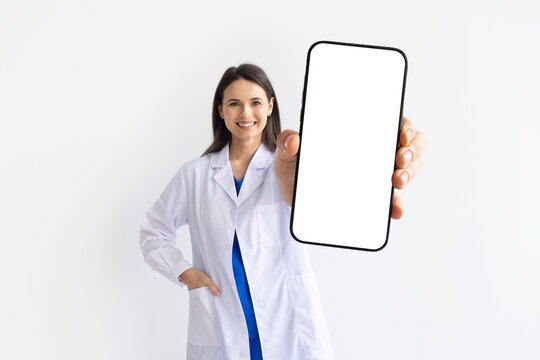 Smiling female doctor in white medical coat presenting smartphone with blank screen on white studio background, concept of digital healthcare and telemedicine