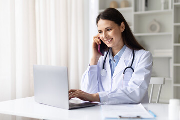 Smiling female doctor in white coat talking on smartphone while working on laptop in clinic office, modern medical communication and online consultation concept