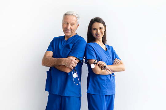 Smiling senior male doctor and young female doctor in blue scrubs standing with arms crossed on white studio background, confident medical team posing together