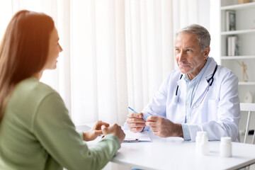 Fototapeta premium Senior male doctor talking with female patient in clinic office, professional consultation and healthcare discussion concept