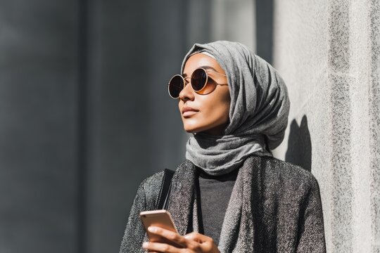 Young woman wearing a hijab and sunglasses holding smartphone, looking away in urban setting