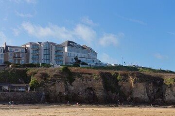 Obraz premium A coastal hotel sits atop a rugged cliff overlooking a sandy beach. The hotel is modern, with balconies and windows facing the ocean in Perranporth - Cornwall - UK