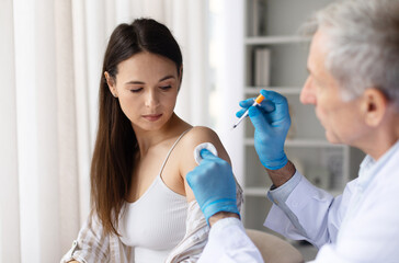 Female patient receiving vaccine injection from senior male doctor wearing gloves in clinic, healthcare immunization and vaccination prevention concept